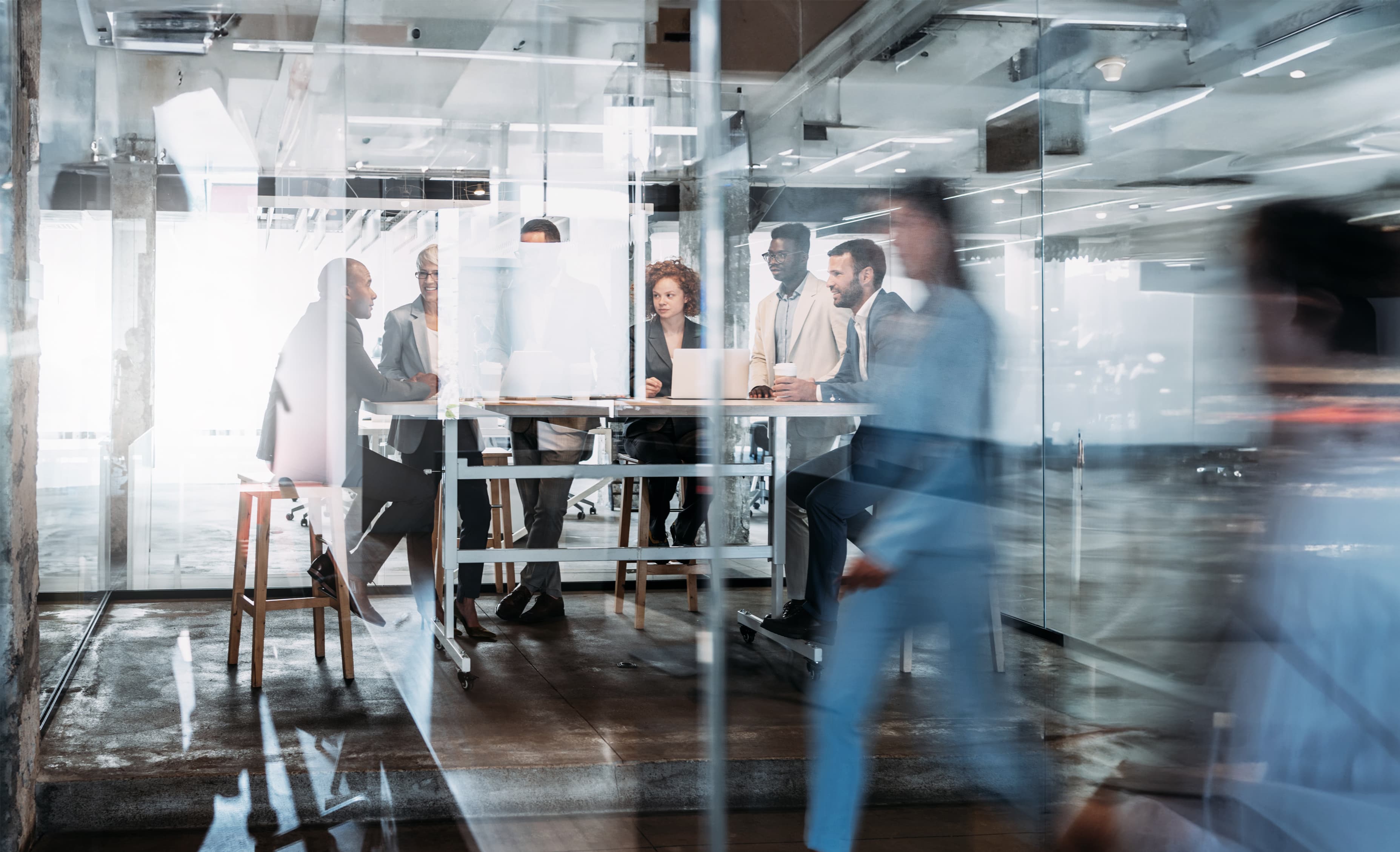 Gruppe von Fachkräften bei einer Besprechung an einem hohen Tisch in einem modernen Büro mit Glaswänden, im Vordergrund verschwommene Bewegung vorbeigehender Personen.