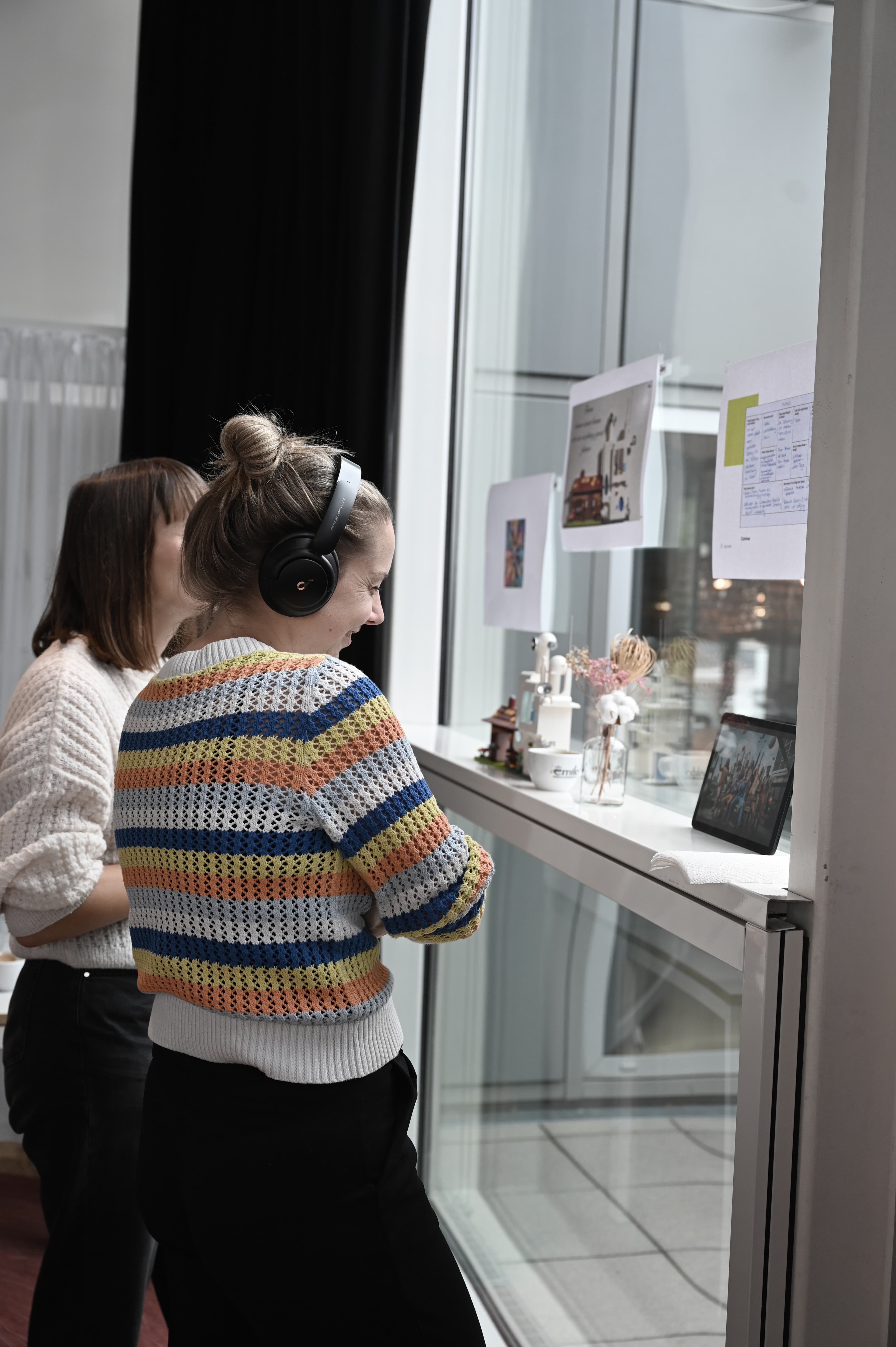 Zwei Frauen stehen an einem Fensterplatz, eine mit Kopfhörern, betrachten ein digitales Tablet und ausgedruckte Bilder an der Scheibe, umgeben von kleinen Dekoelementen.