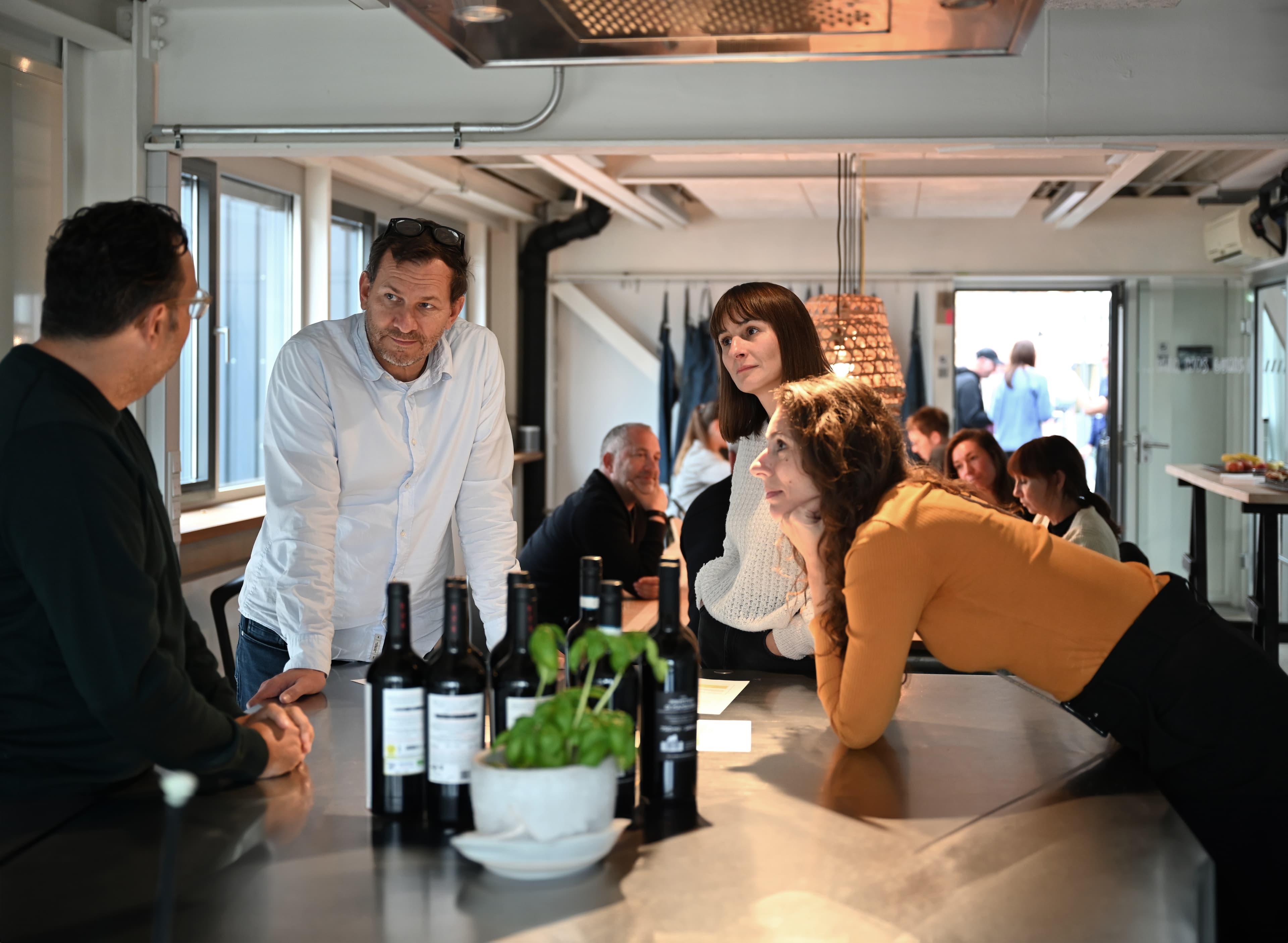 Gruppe von Personen im Gespräch an einer Kücheninsel in einem modernen Büroraum, mit Weinflaschen und einem Basilikumtopf auf der Arbeitsfläche.