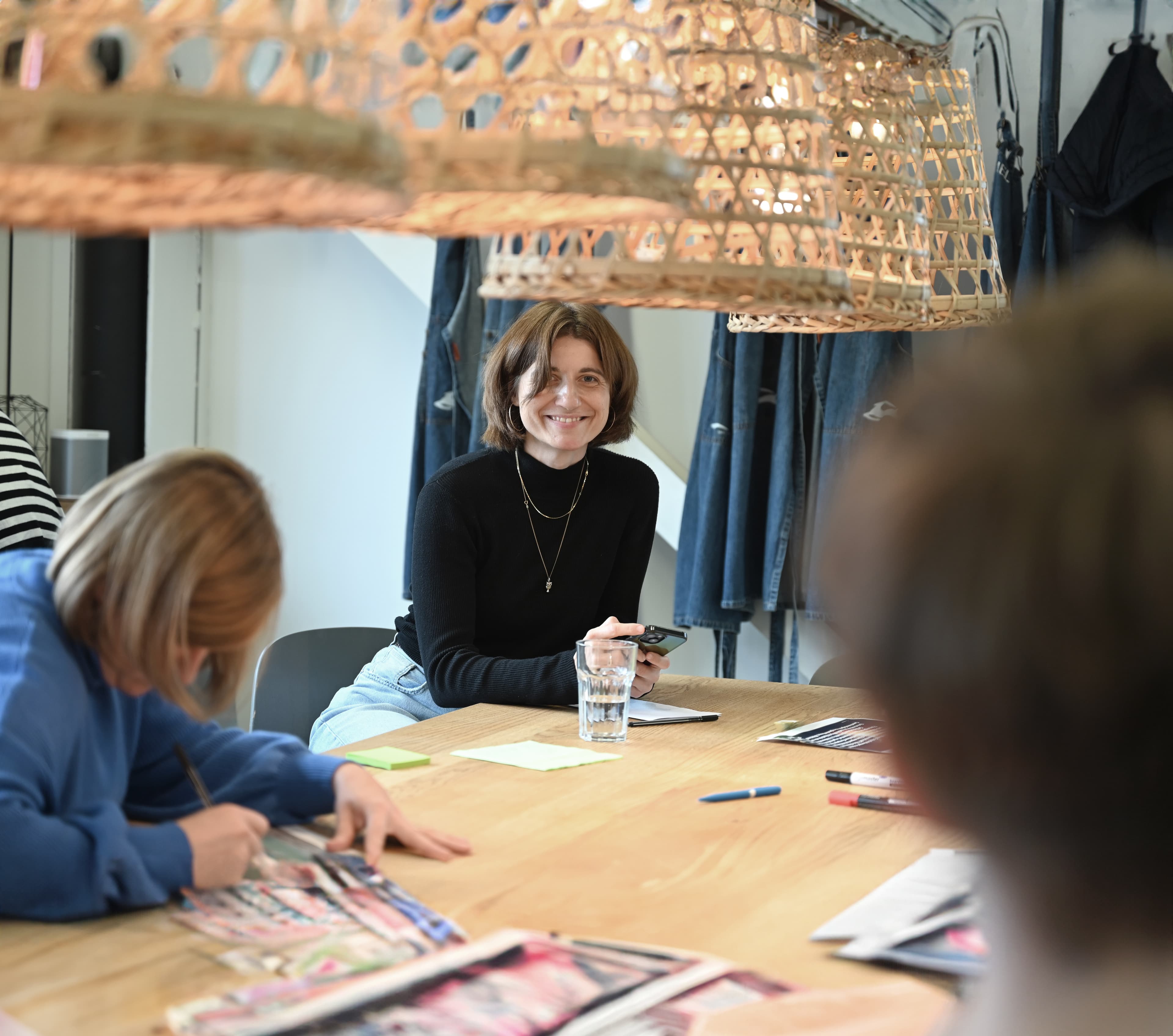 Frau lächelt und hält ein Handy in der Hand, während sie mit anderen an einem kreativen Workshoptisch unter geflochtenen Hängelampen sitzt.
