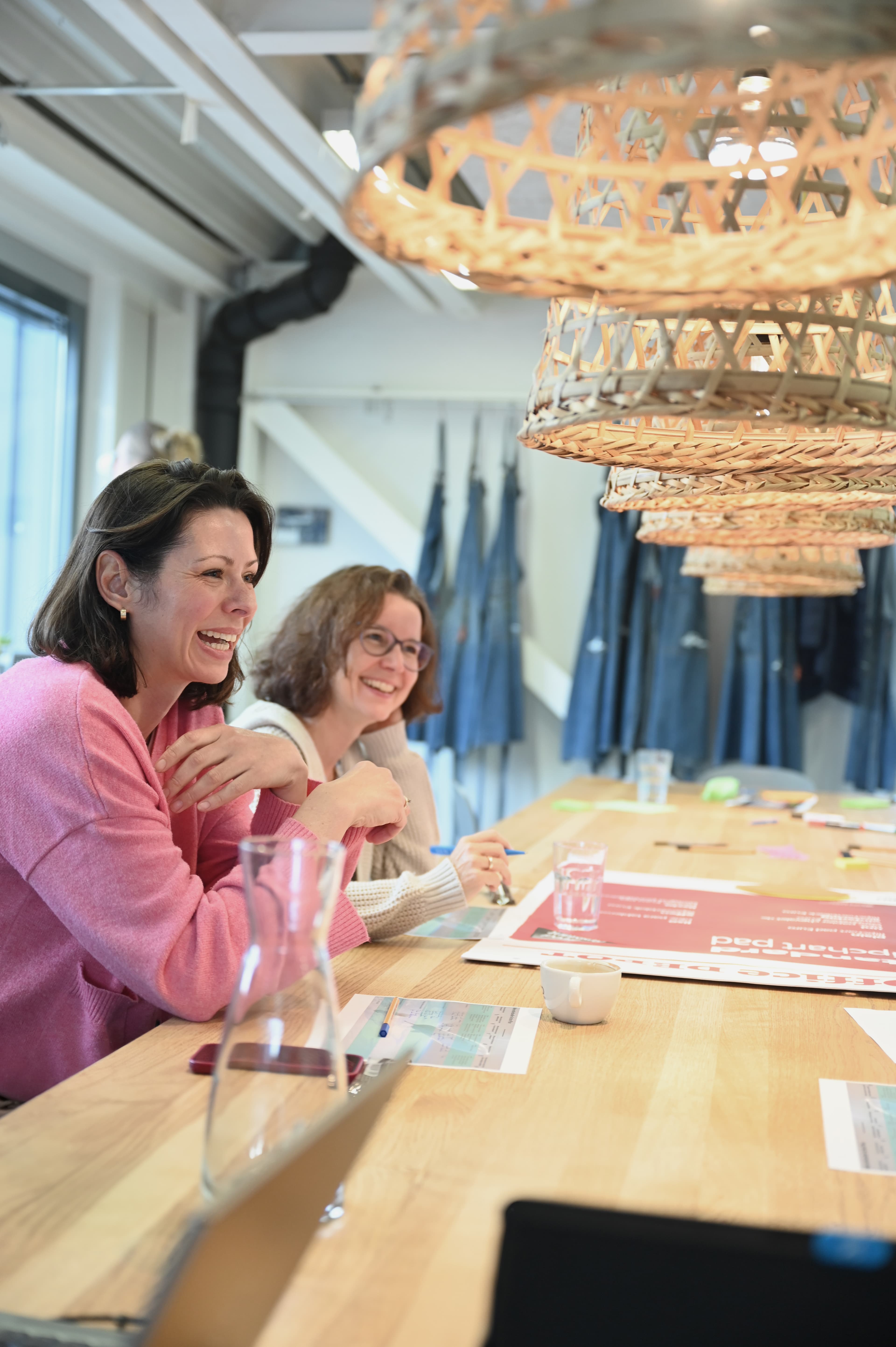 Zwei Frauen lachen und arbeiten an einer kreativen Sitzung an einem langen Holztisch, umgeben von Unterlagen, Markern und geflochtenen Hängelampen.