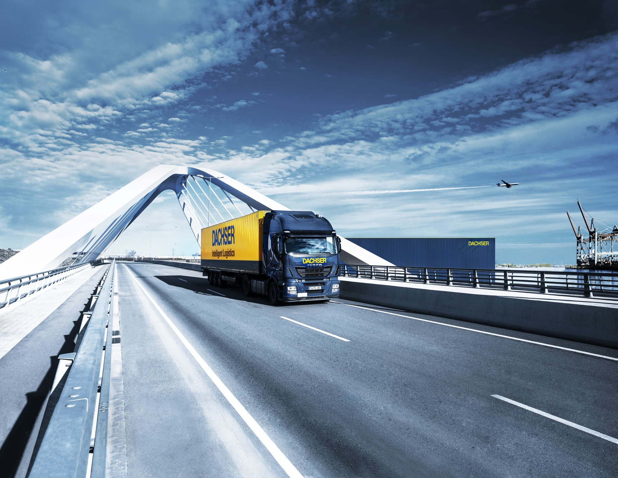 DACHSER-Logistik-Lkw überquert eine moderne weiße Brücke mit Lagerhalle und Flugzeug im Hintergrund unter blauem Himmel.
