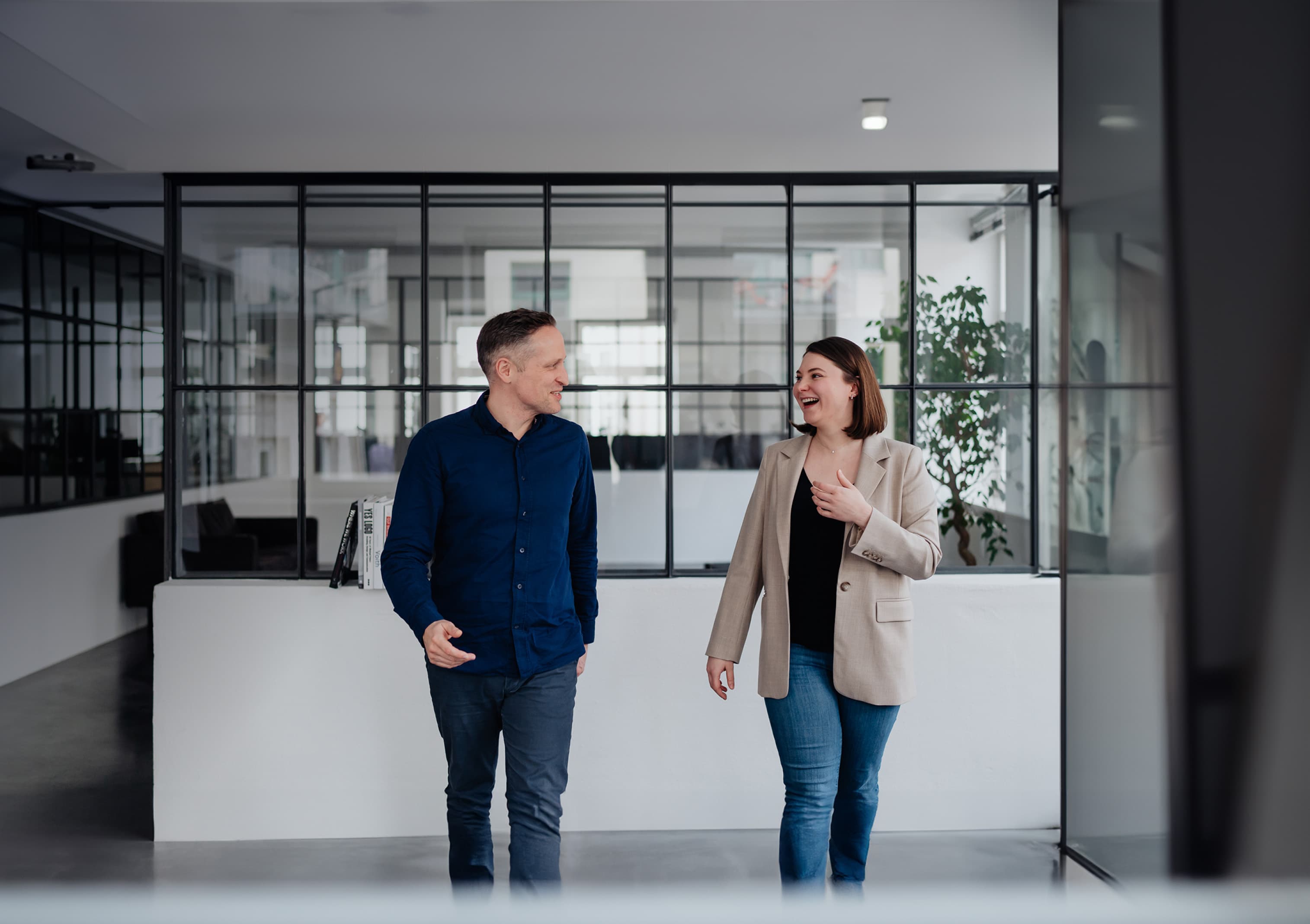 A Man and a woman walking through a modern office hallway engaged in a lively conversation.
