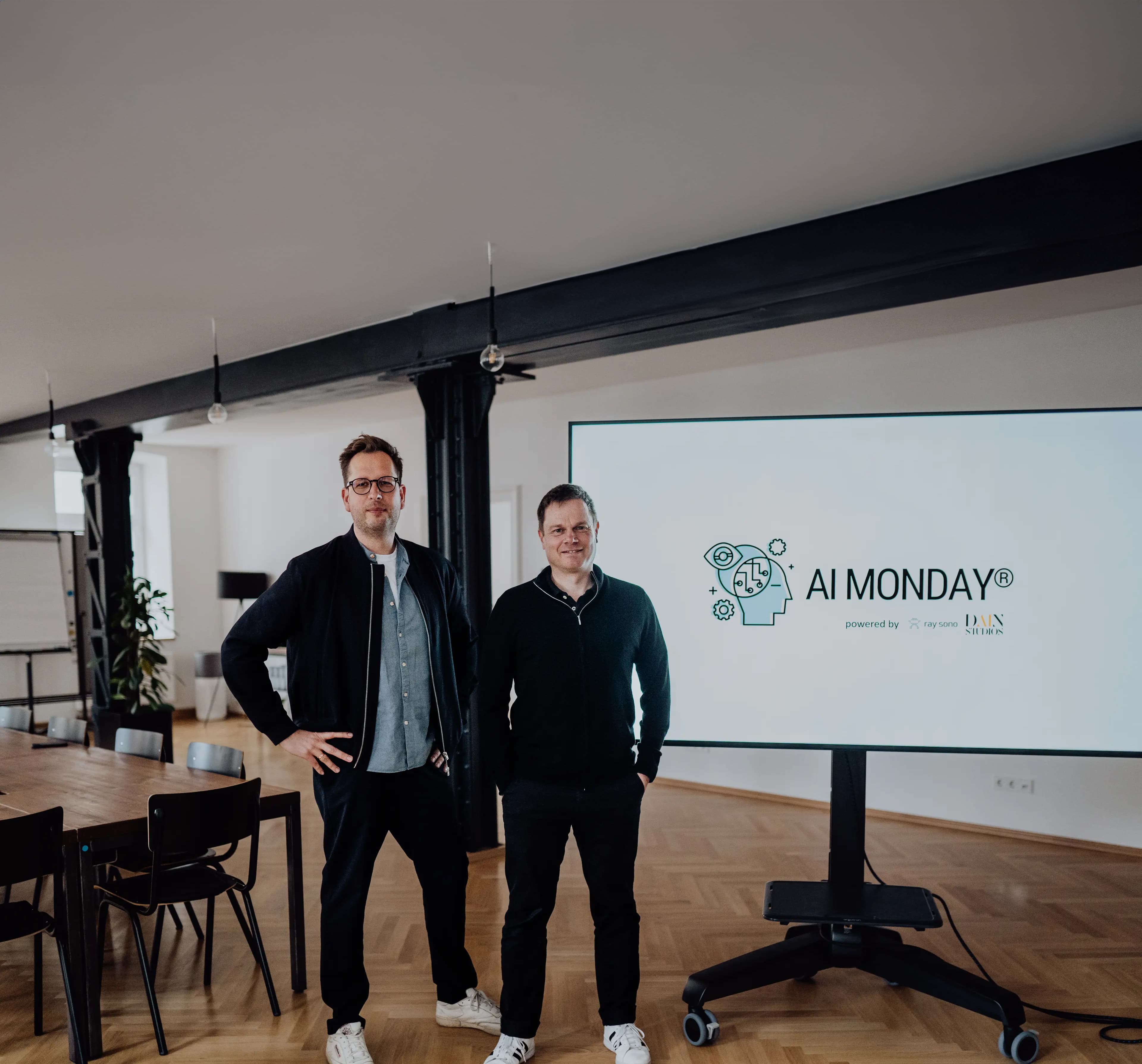 Zwei Männer stehen neben einem großen Bildschirm mit dem Logo und Branding von AI MONDAY® in einem modernen Besprechungsraum.
