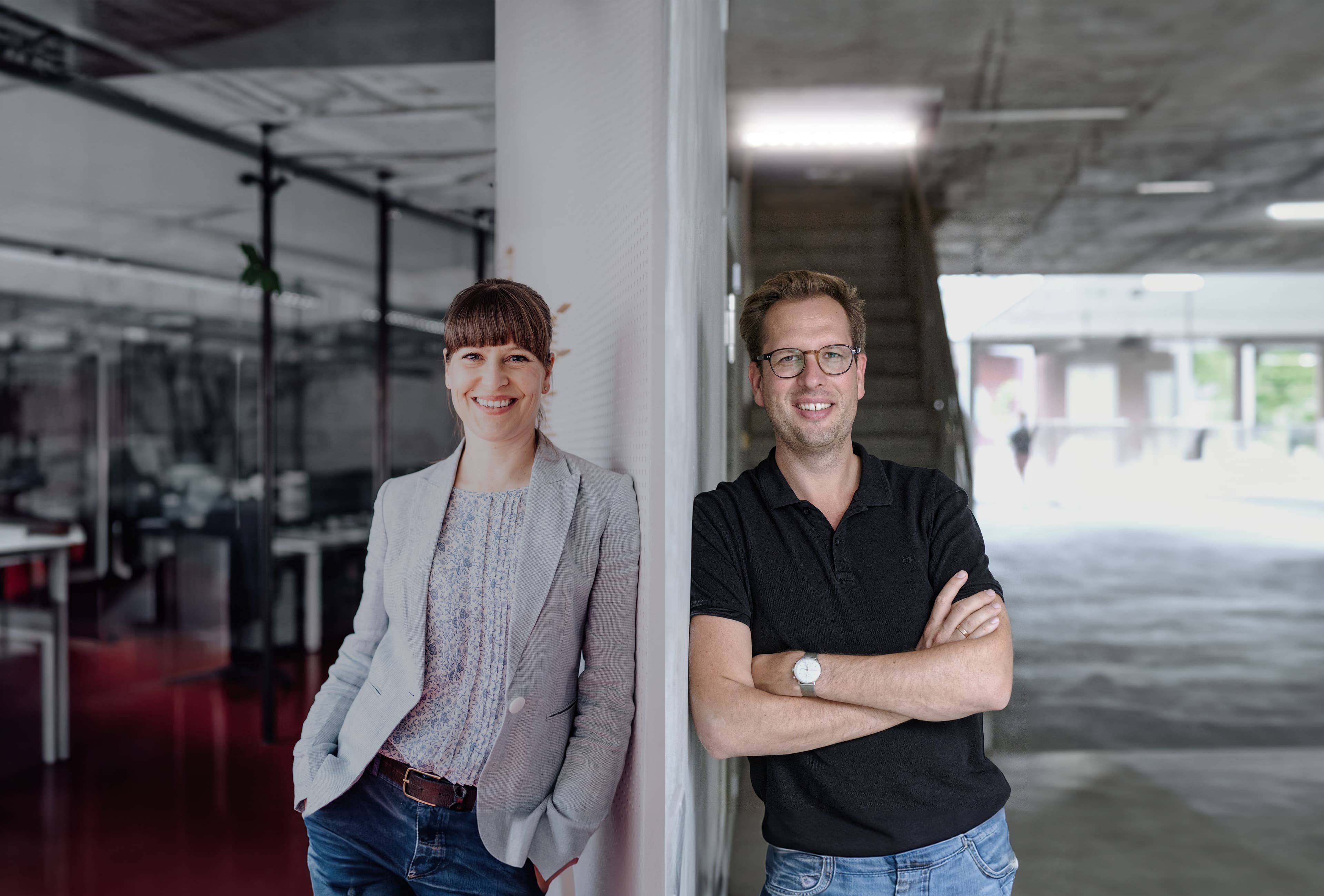 Die Vorstandsmitglieder Ilona Kühn und Sebastian Krüger lehnen entspannt an einer Wand in einem modernen Bürogebäude mit Glaswänden und offenen Arbeitsbereichen.