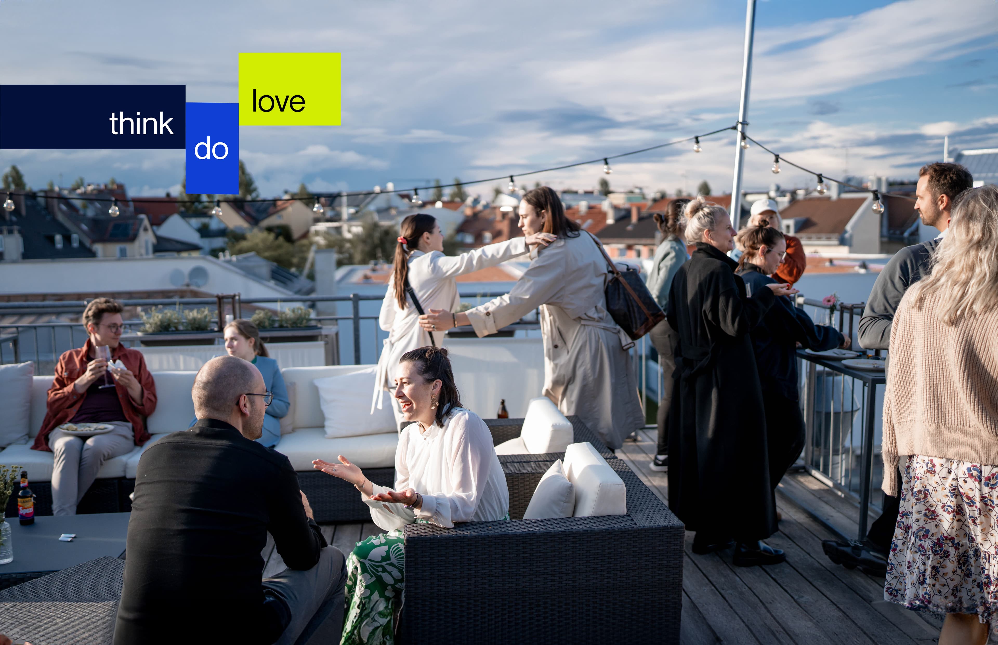 Menschen unterhalten sich auf einer Dachterrasse mit Blick über die Stadt und teils bewölktem Himmel, mit dem Ray Sono Slogan think do love in farbigen Blöcken überlagert.