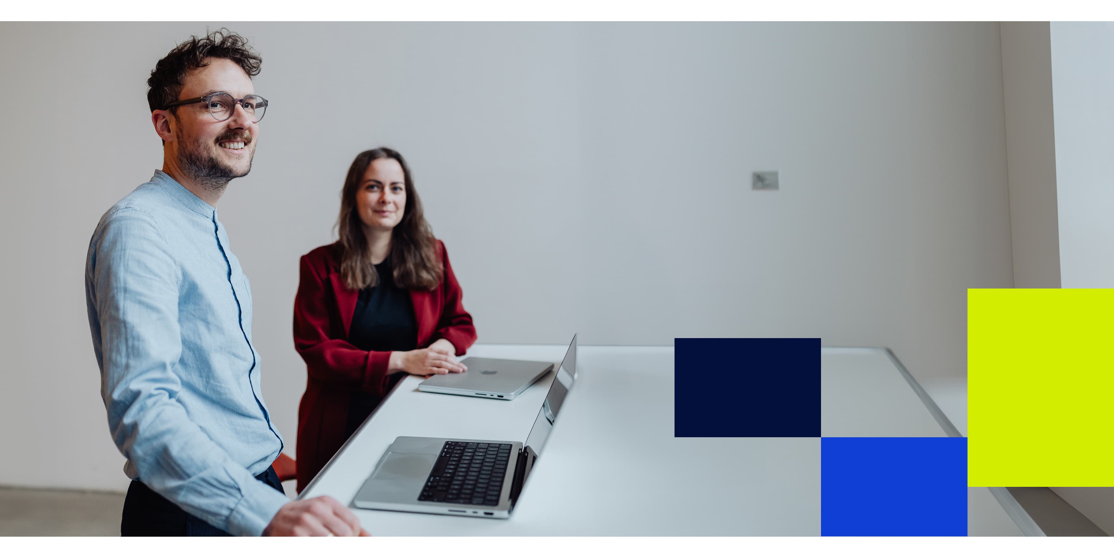 Two colleagues at a white desk with laptops, smiling in a bright room, accompanied by bold blue and yellow graphic blocks on the right.