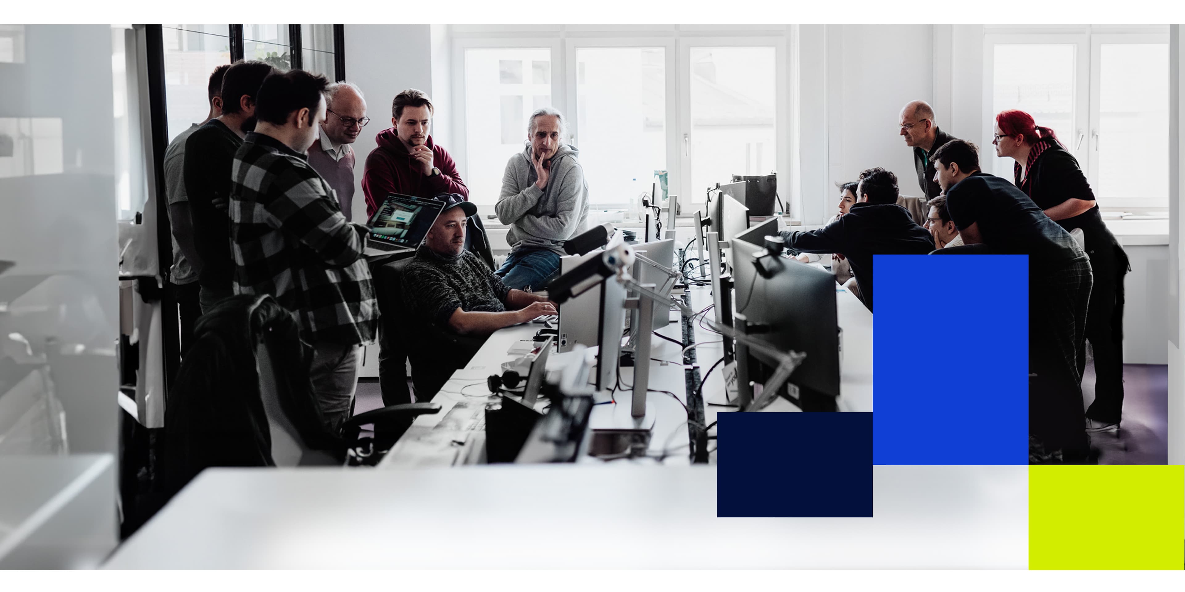 Team gathered around computer screens in a modern office, actively discussing and collaborating, with bold blue and yellow graphic elements on the right.