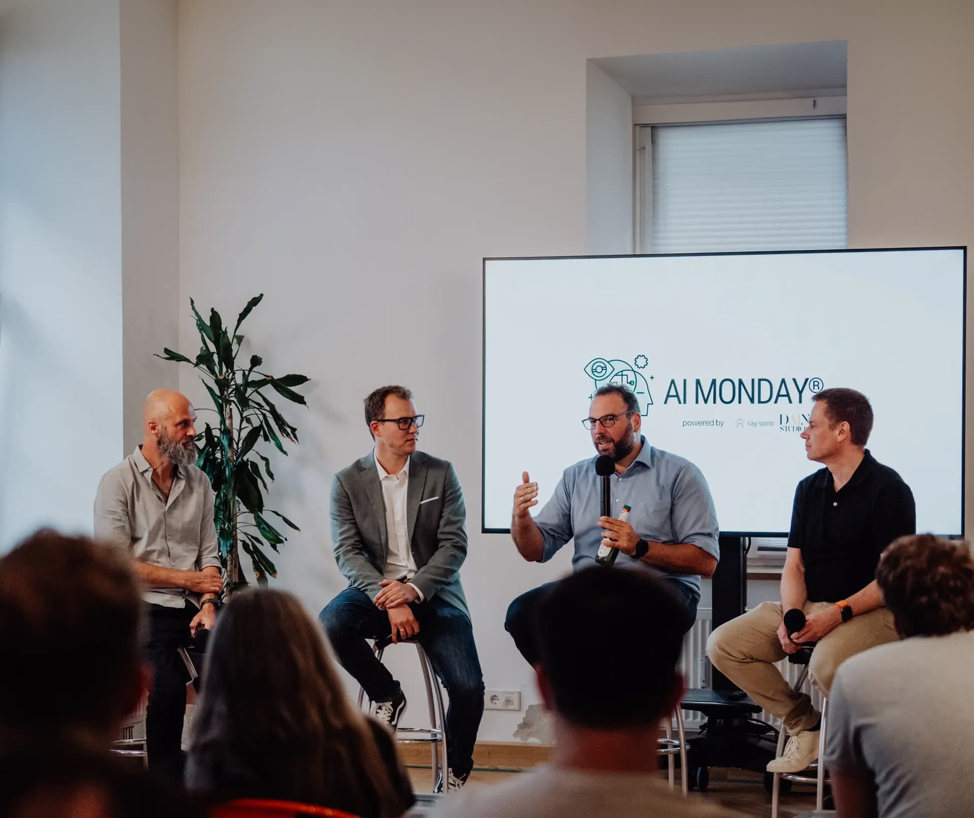Vier Männer diskutieren in einer Podiumsrunde bei einem AI MONDAY®-Event, sitzend vor einem Bildschirm mit dem Event-Branding, während das Publikum zuhört.