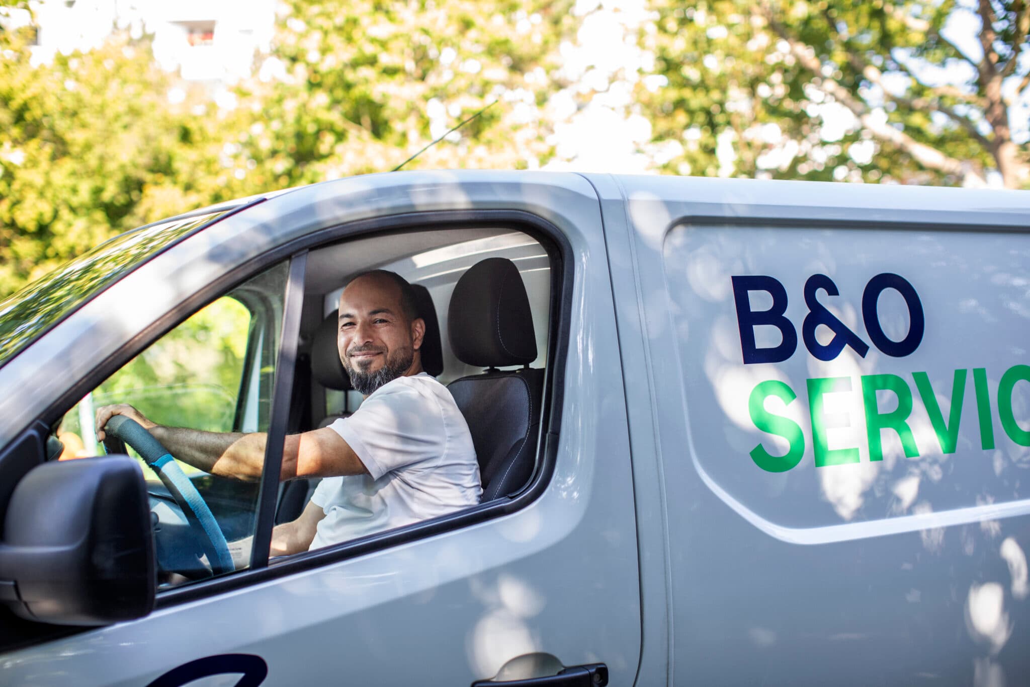 B&O Service worker with arms crossed in blue sweater in a hallway with pipes on ceiling. Maintenance or service in the background.