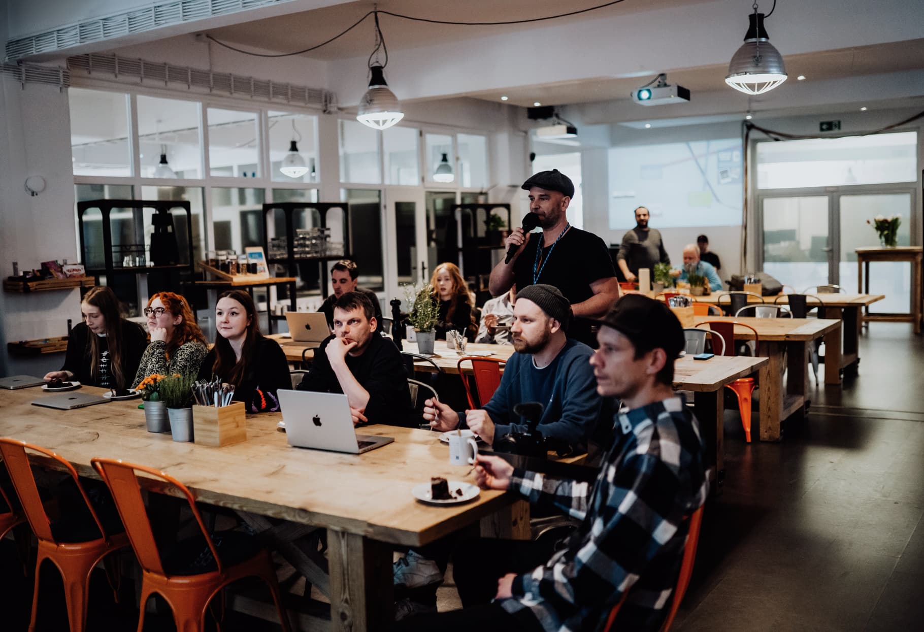 Personen sitzen an großen Holztischen mit Laptops und Kaffee und folgen aufmerksam einer Präsentation in einem hellen, offenen Arbeitsraum.