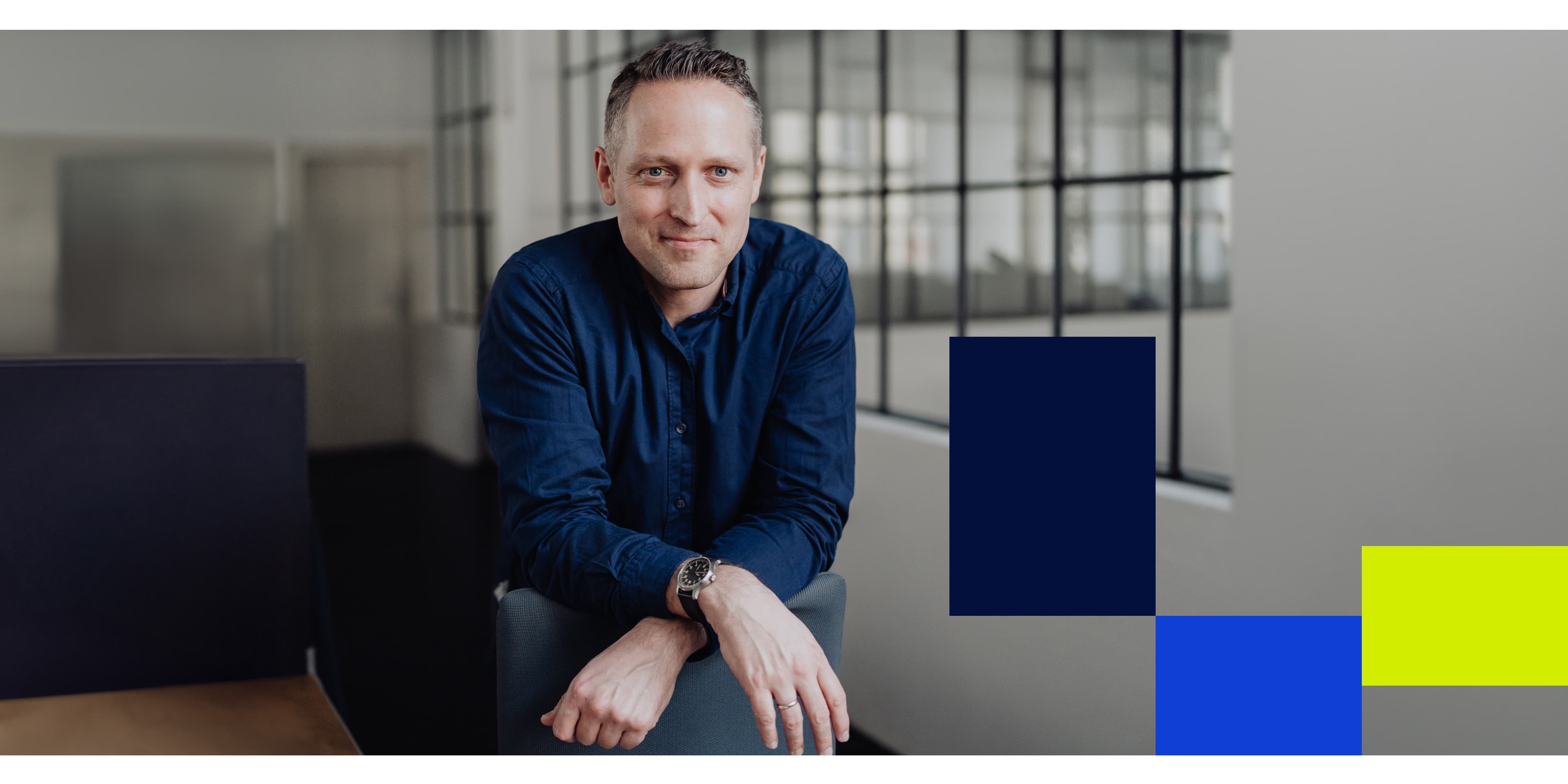 Man in a dark blue shirt leaning casually on a chair in a modern office space, with graphic blocks in navy, blue, and yellow on the right.