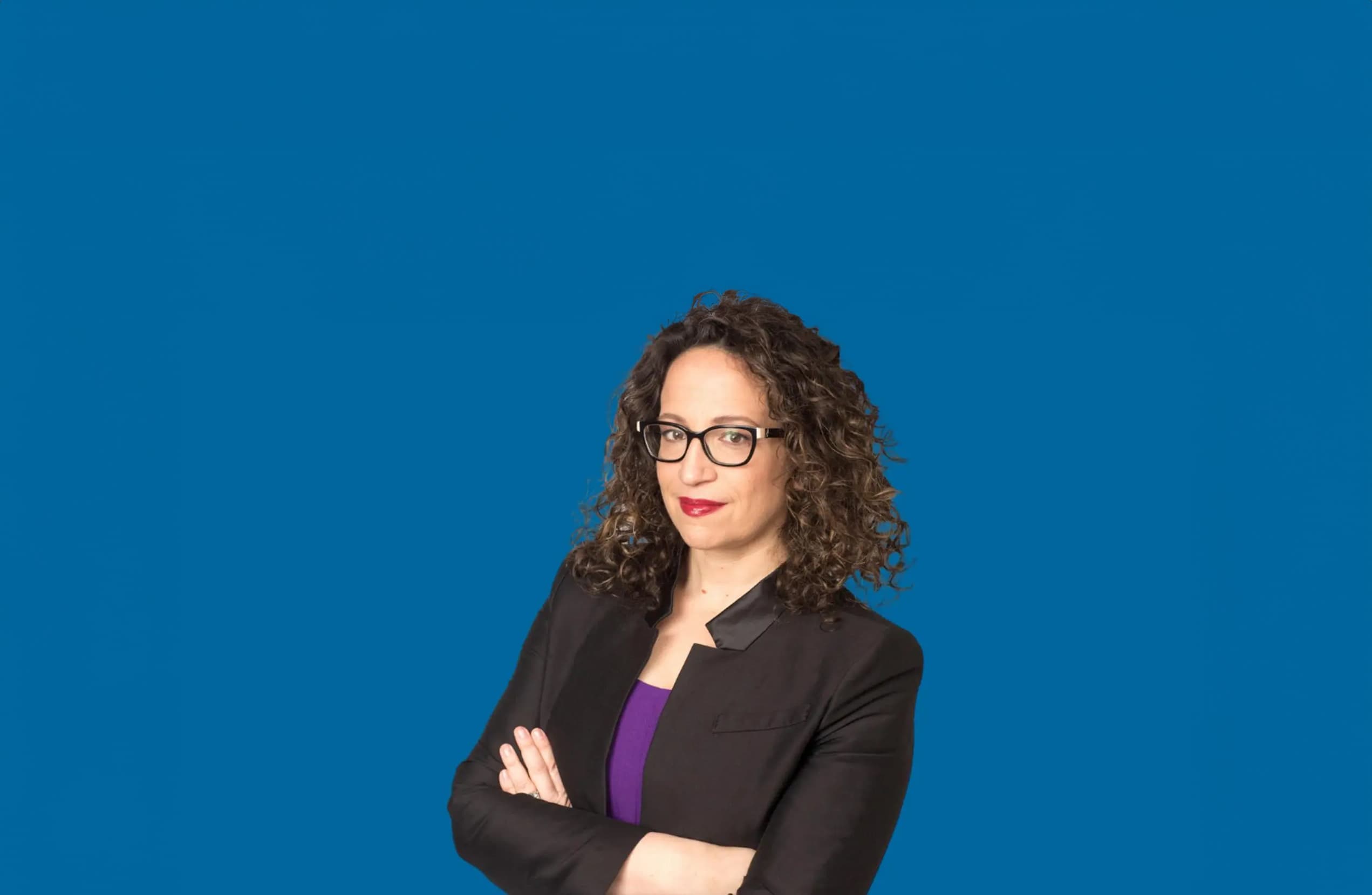 Selbstbewusste Frau mit lockigem Haar und Brille in schwarzem Blazer steht mit verschränkten Armen vor blauem Hintergrund.